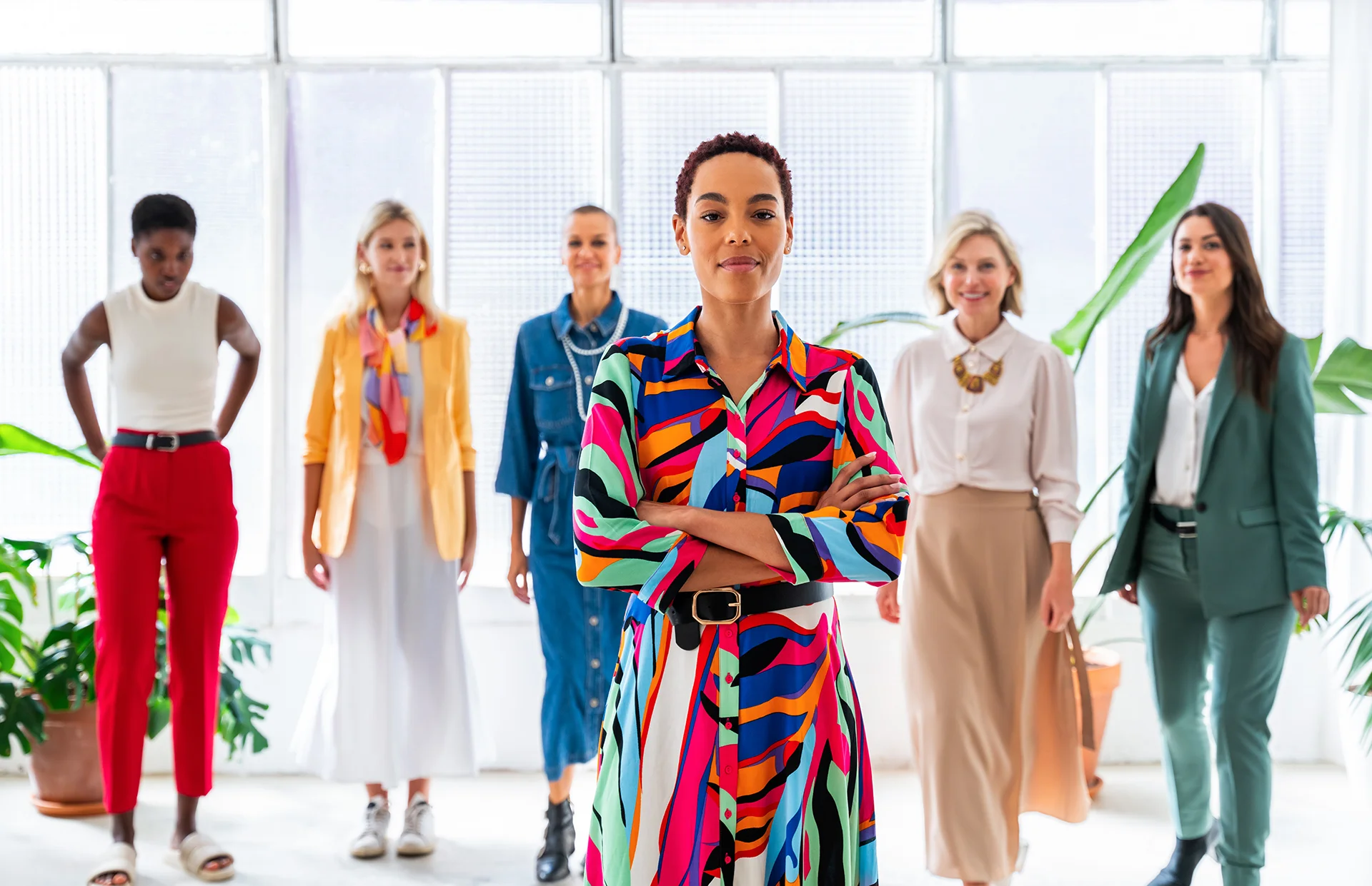 A diverse group of six businesswomen stand together in a brightly lit office, with a woman in a colorful patterned dress in the foreground and five colleagues posing in the background.